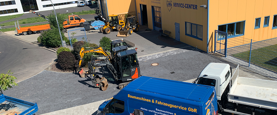 Blick von oben auf den LöWa-Hof: gepflasterter Platz mit Radlader, Minibaggern und dem blauen Servicetransporter vor der Halle.