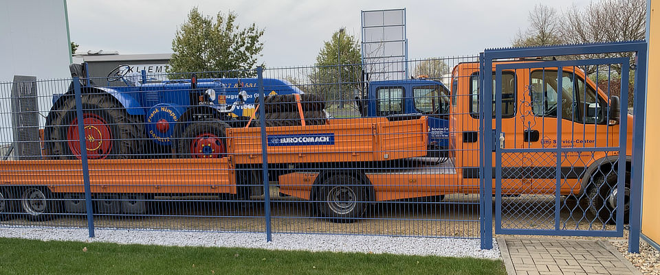 Orange Transportfahrzeuge von LöWa hinter dem verzinkten Metallzaun des Betriebsgeländes.